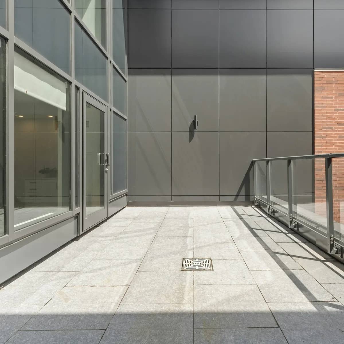 An empty condo balcony paved in pale stone, with a glass-and-steel railing and a floor drain, in afternoon light.