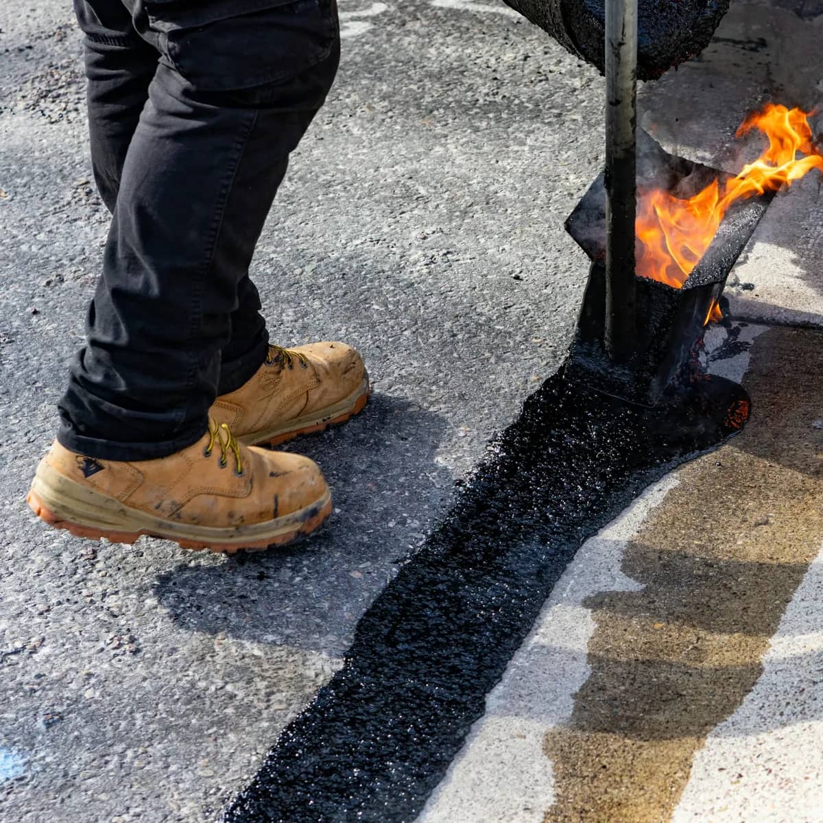 A worker in boots running a torch-heated applicator along a joint, laying a line of hot asphalt sealant.