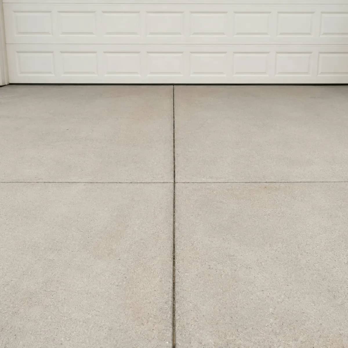 An evenly coloured concrete driveway with saw-cut control joints leading up to a white garage door.