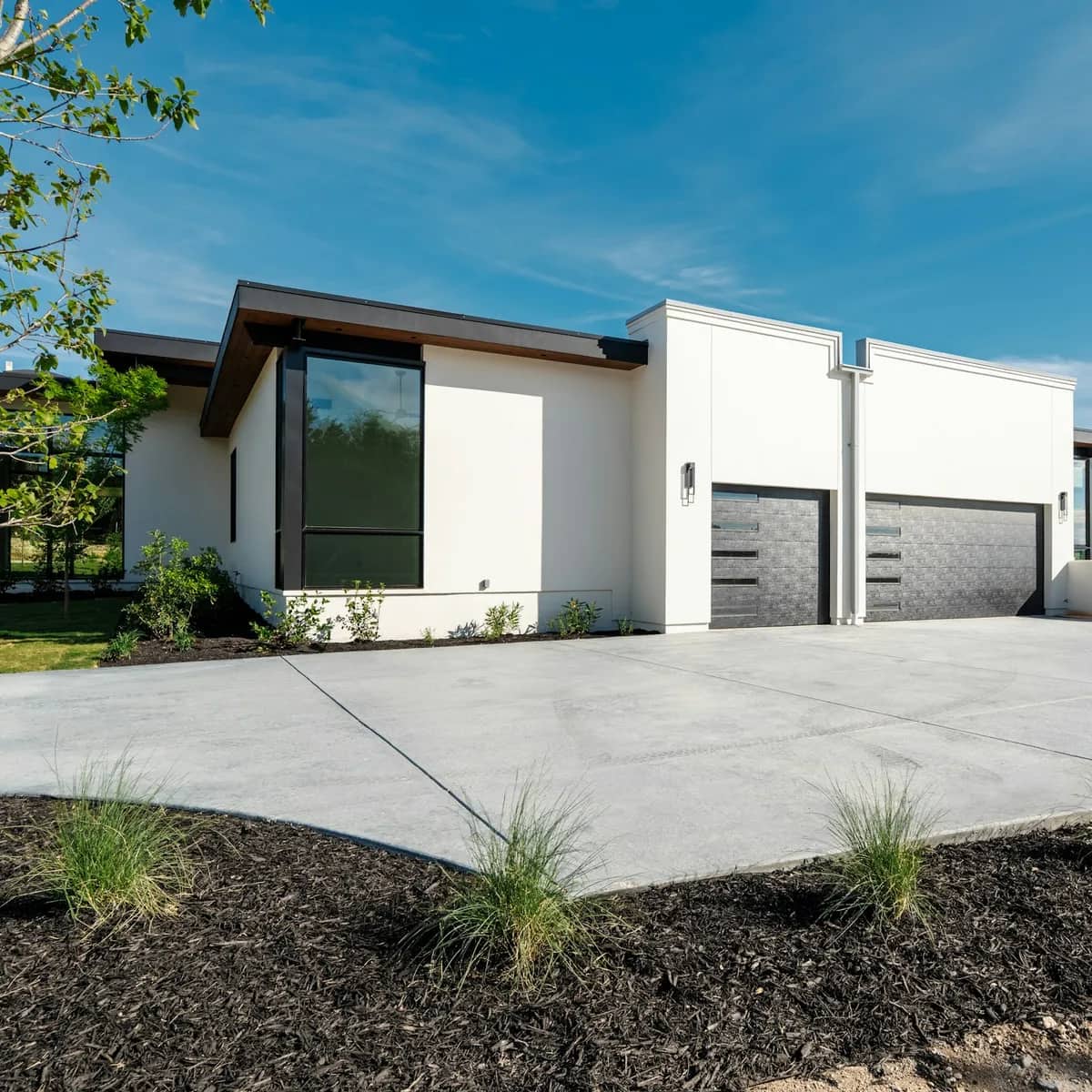 A wide, pale concrete driveway in front of a modern white house with two dark garage doors.