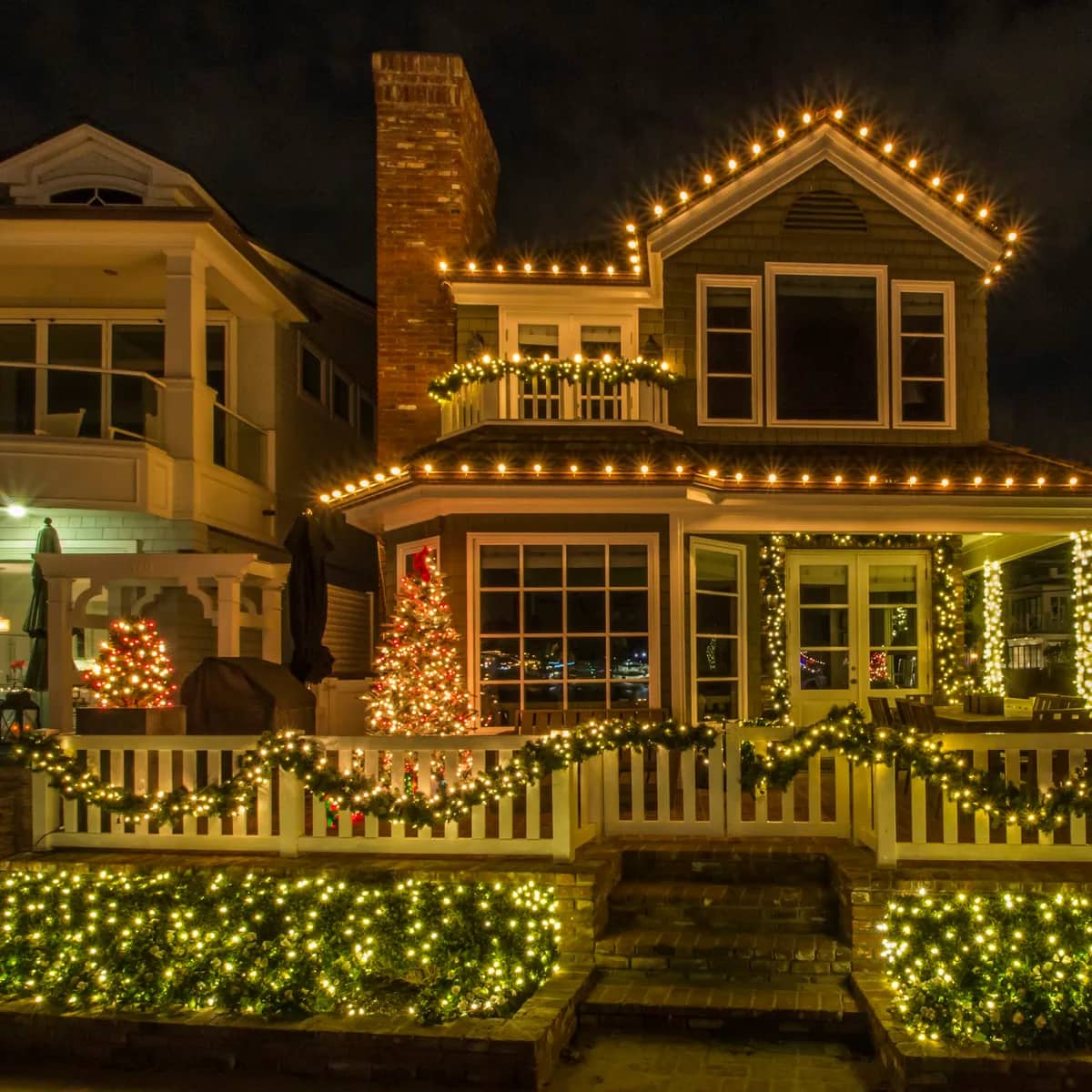 A house at night outlined in warm white bulbs along every roofline, with lit garlands on the fence.