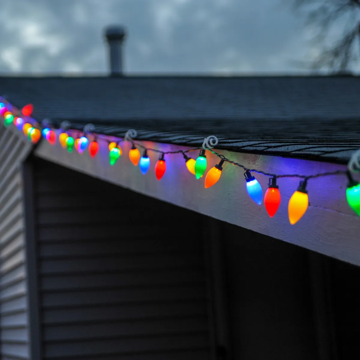 Large multicoloured C9 bulbs clipped along the eave of a grey-sided house at dusk.