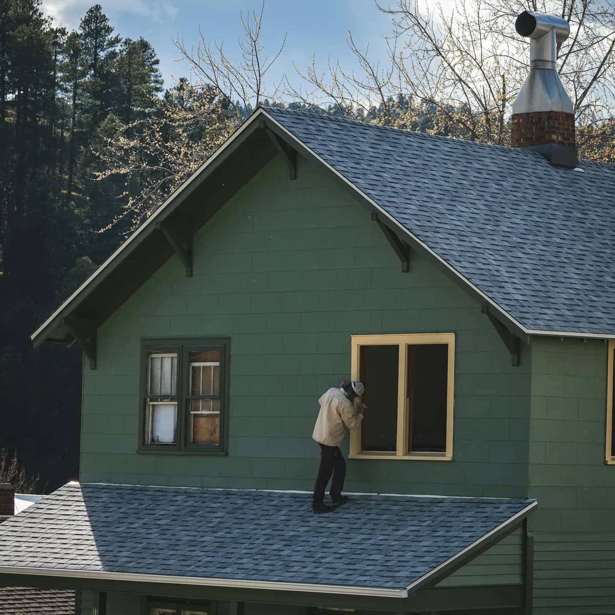 A painter standing on a porch roof, working on the yellow window trim of a green shingled house.