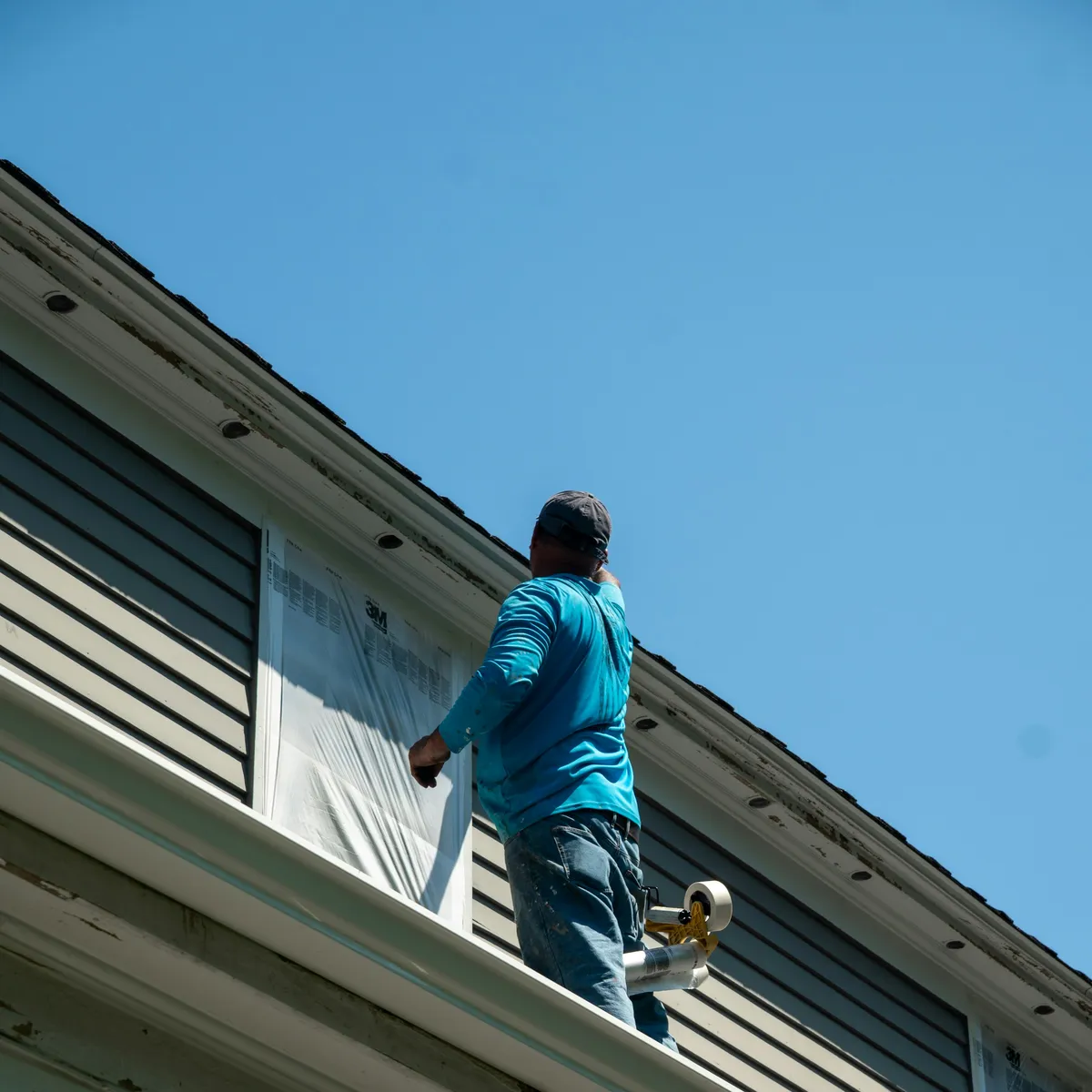 A worker on the roof edge taping plastic sheeting over a window on grey lap siding.