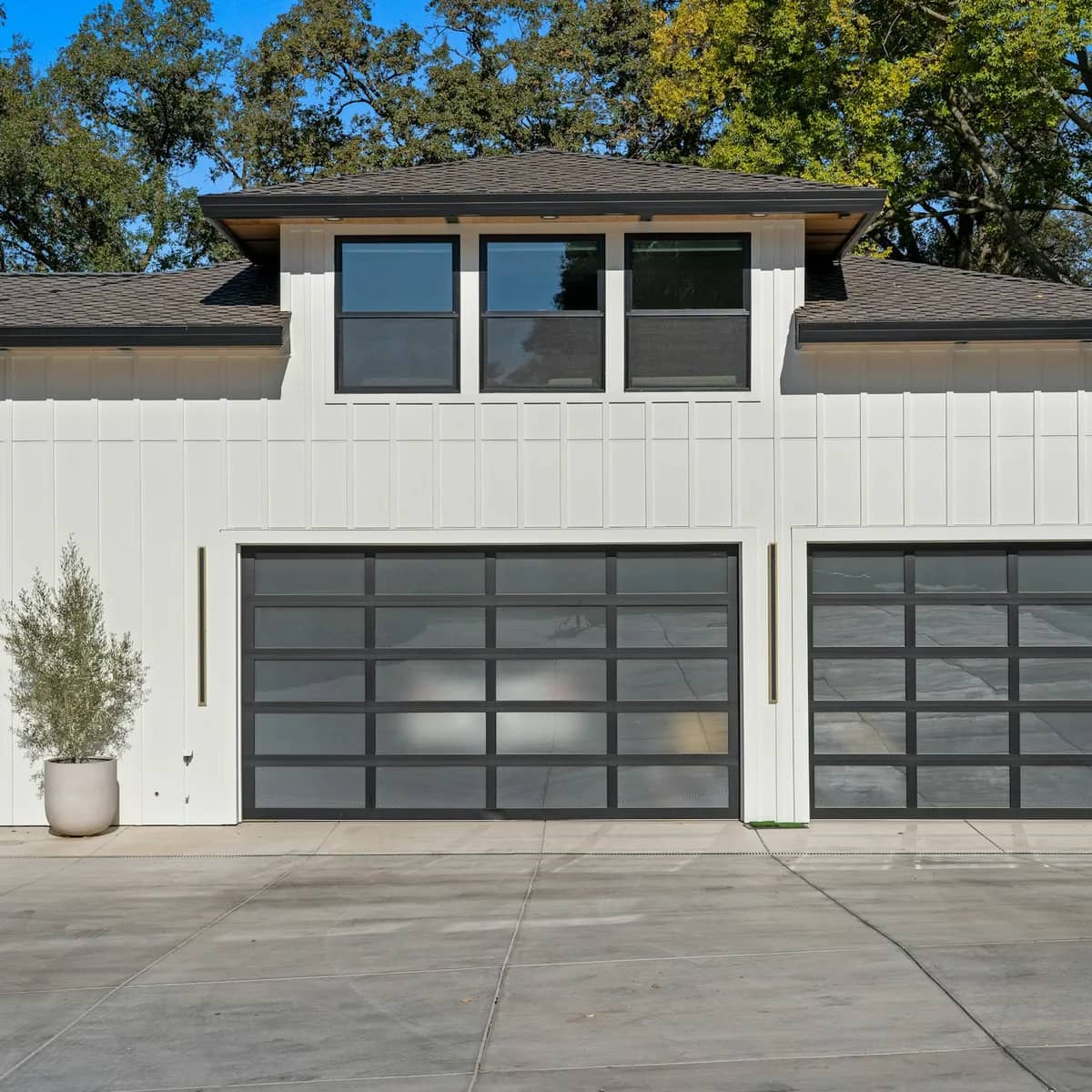 A white board-and-batten house with two dark glass garage doors and potted olive trees, the siding clean.