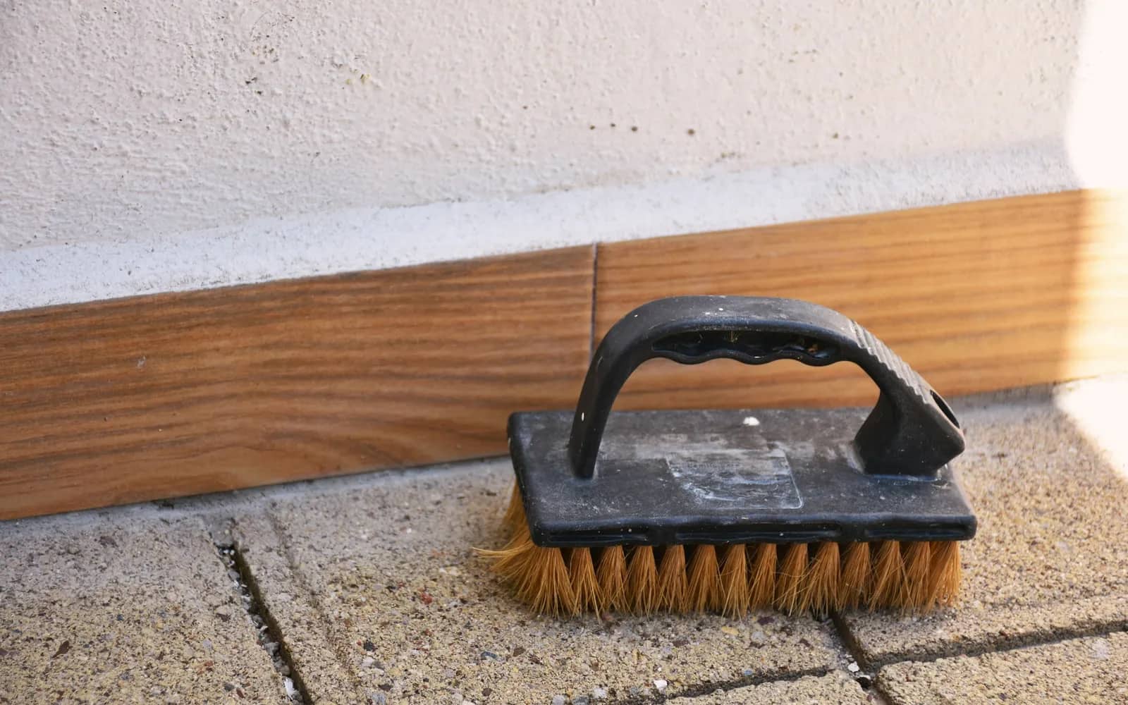 Balcony cleaning: a stiff hand scrubbing brush resting on concrete pavers against a white wall with a timber skirting board.