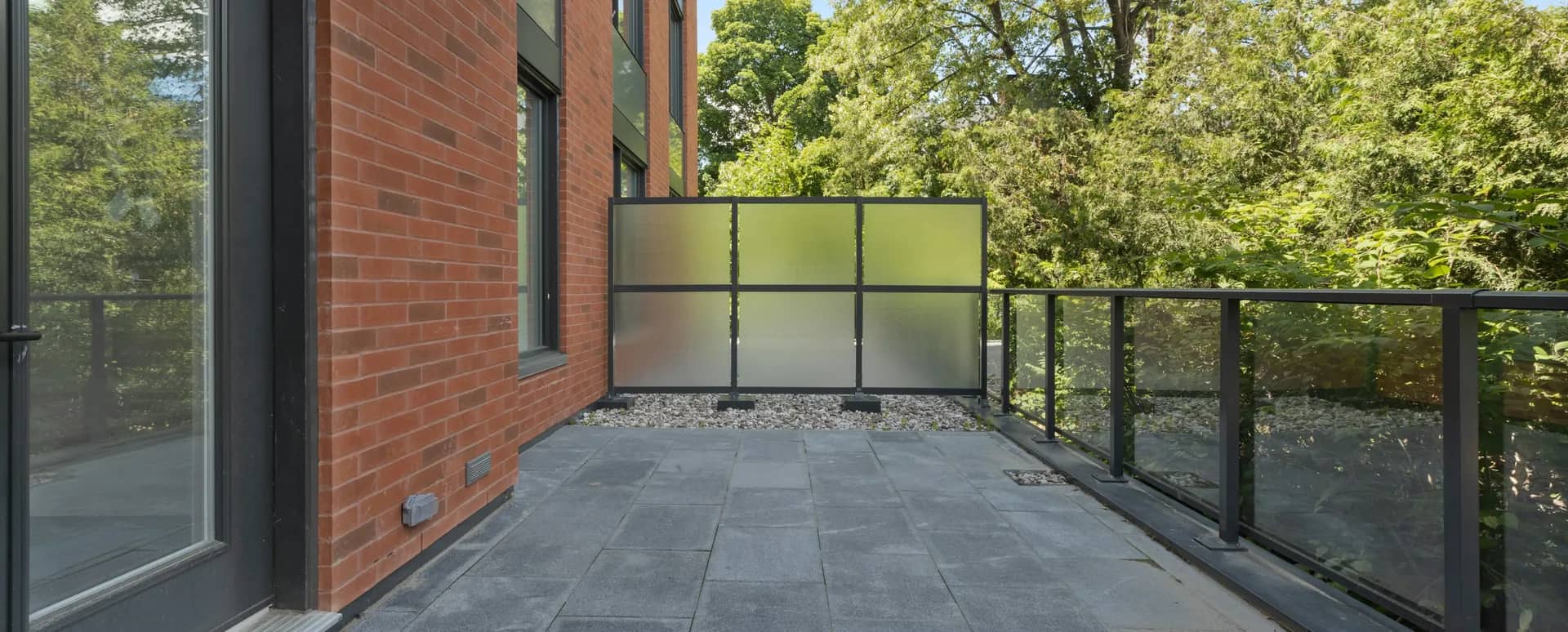 A clean paved balcony beside a brick wall, a frosted privacy screen and a black-framed glass railing, with deciduous trees beyond.