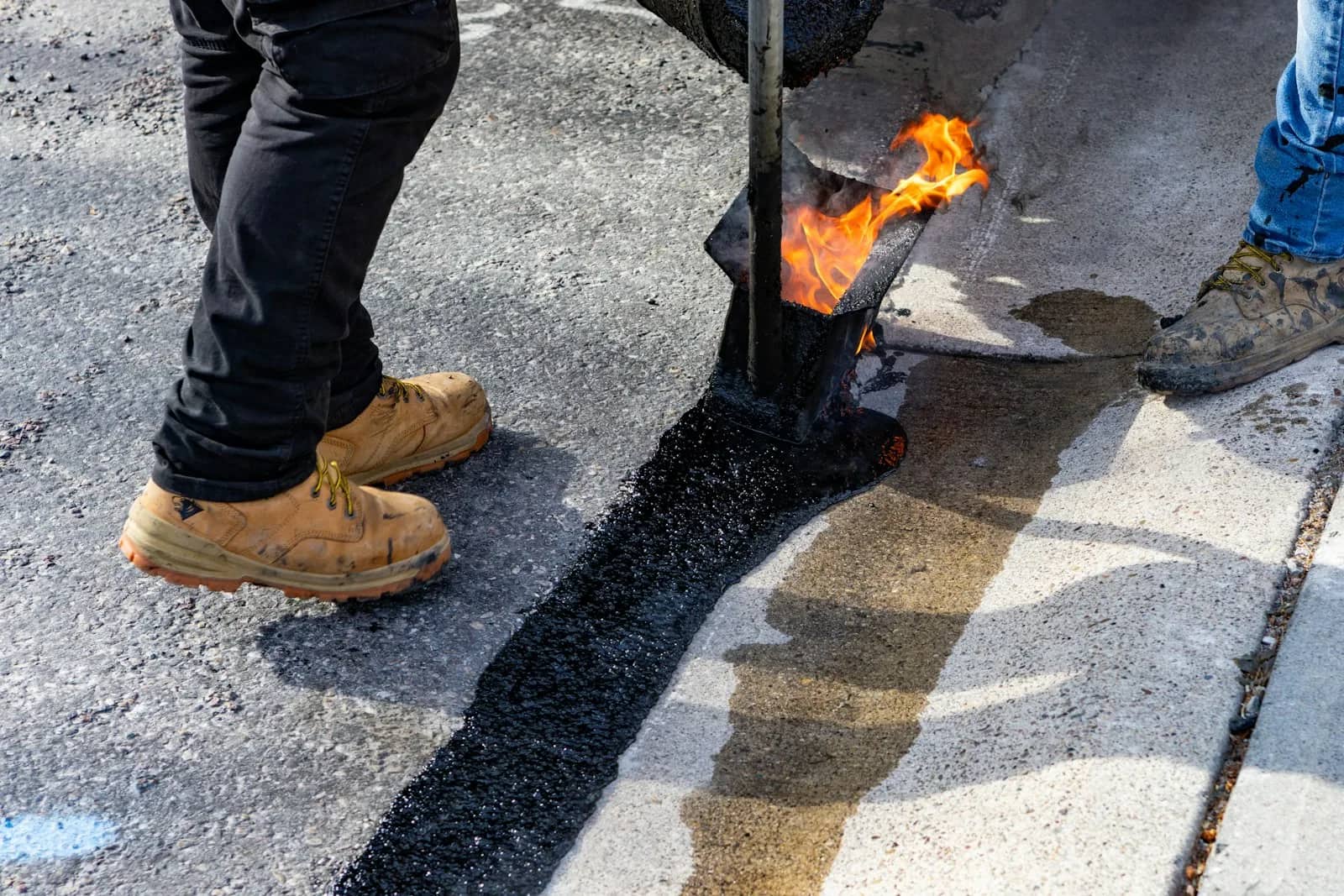 Concrete and asphalt sealing: an operator laying a bead of hot-pour sealant along a joint with a torch applicator, the finished bead running black across the slab behind their boots.