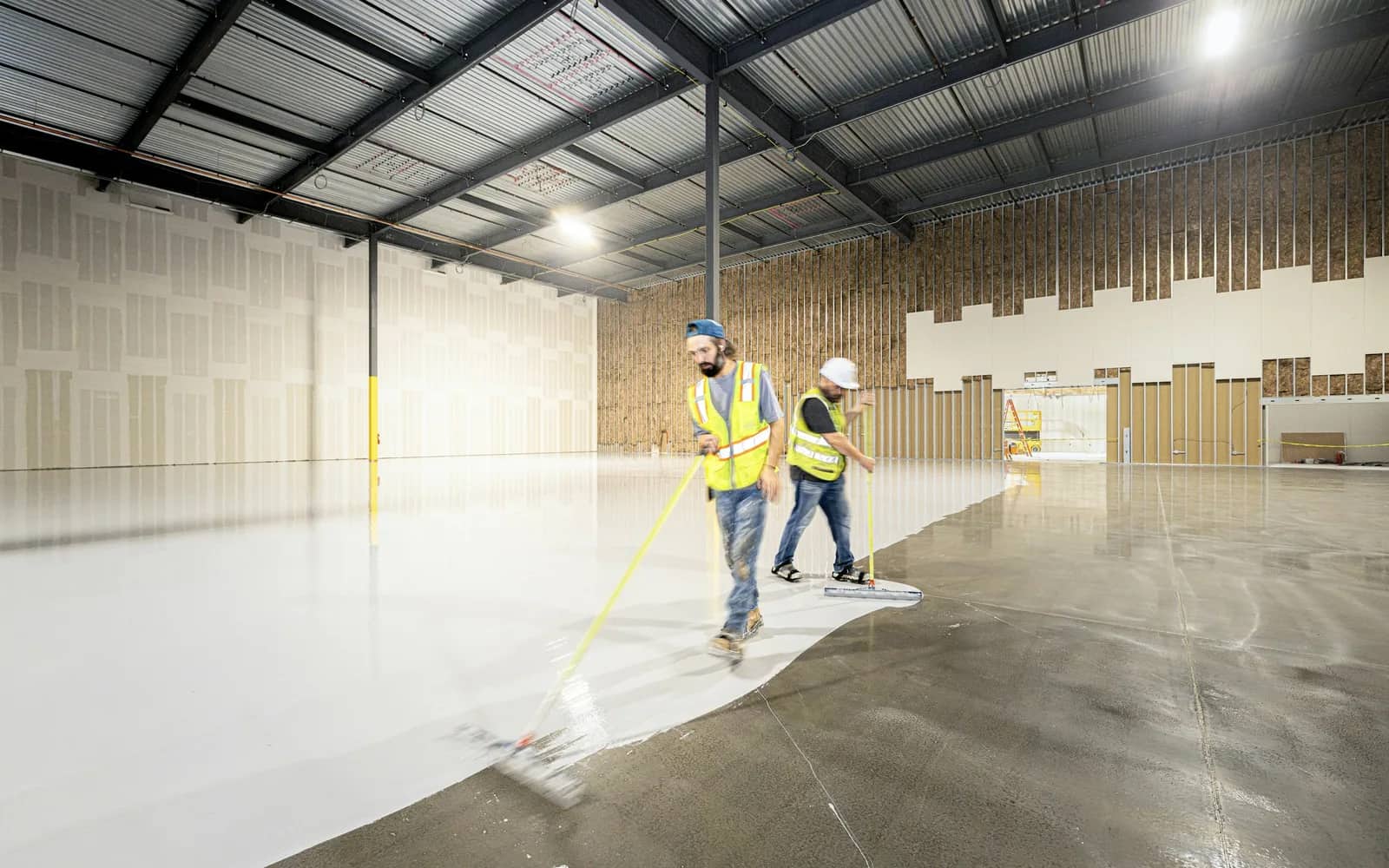 Sealing: two workers in hi-vis vests squeegeeing a pale coating across a bare concrete warehouse floor, the coated half glossy against the grey slab.