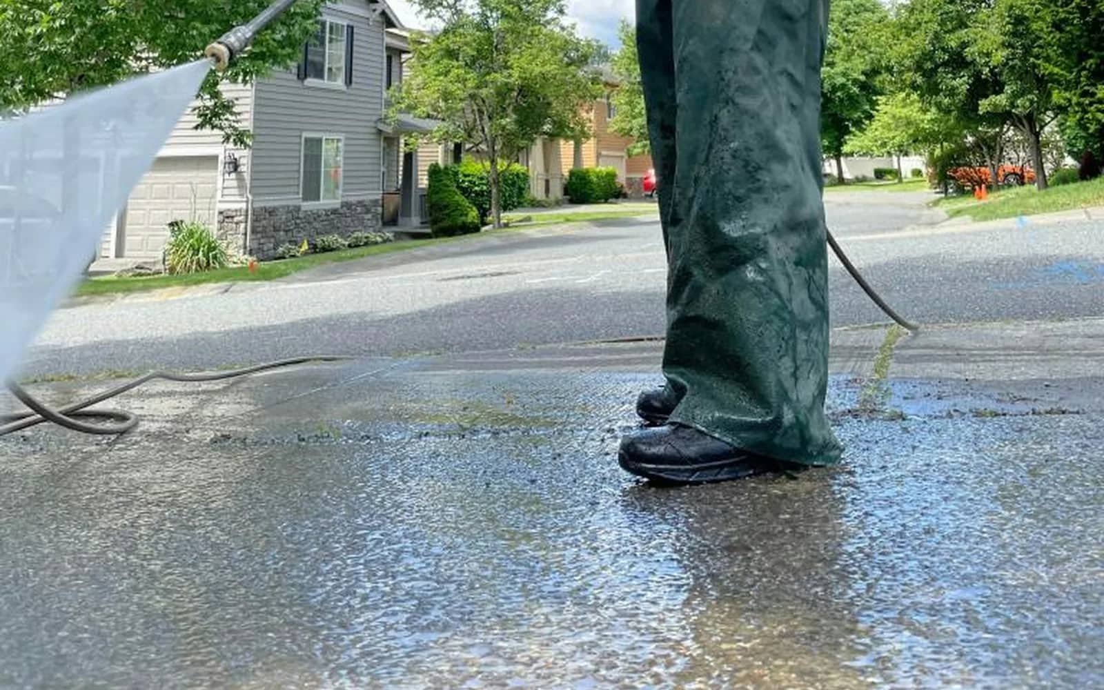 Sealing prep: a worker in waterproof bib trousers pressure washing a concrete driveway, the wet slab darkening where the spray has passed.