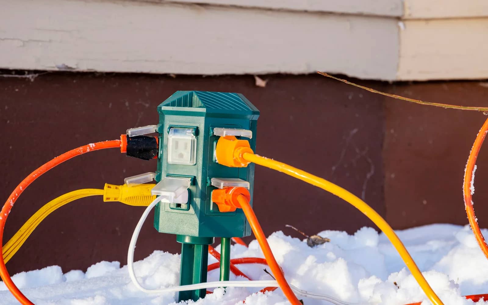 Holiday lights: an outdoor power stake pushed into the snow beside a house, orange and yellow extension cords plugged into its weatherproof outlets.