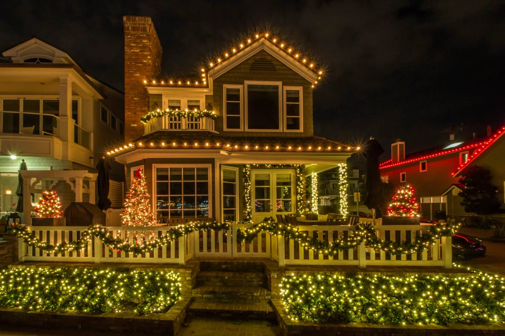 A large house seen from the street at night with its eaves, gables, porch and garden all lit in warm white, the whole frontage reading as one scheme.