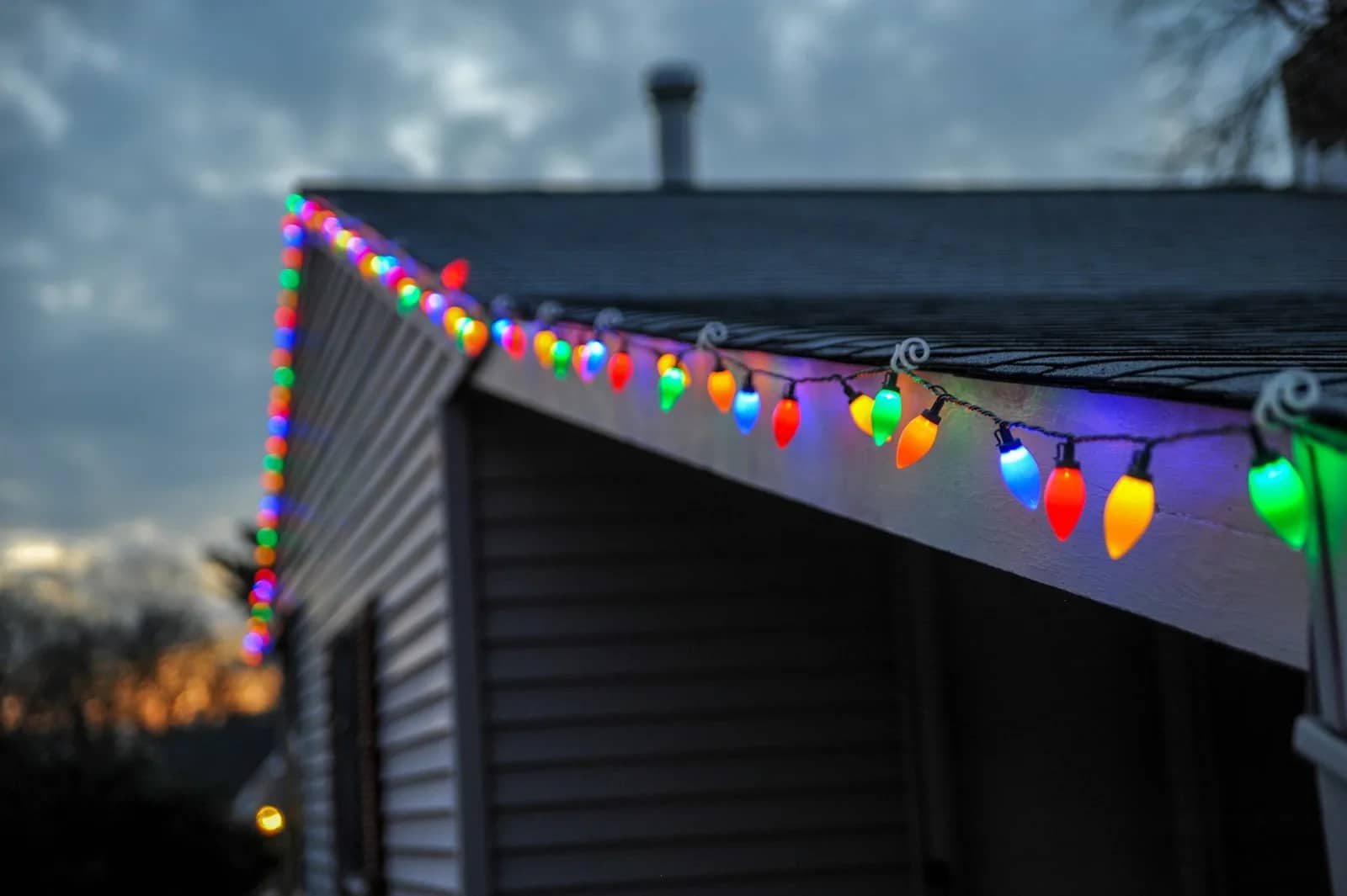 Holiday light installation: a run of large coloured bulbs clipped along the eave and gable of a house at dusk, the spacing even the whole length of the line.