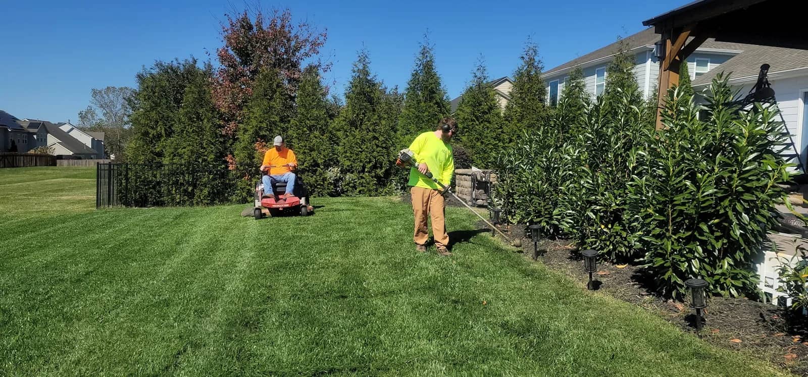 Landscaping and lawn care: two groundskeepers working a lawn together, one riding a mower along a cut stripe while the other trims the edge of the planted bed beside it.