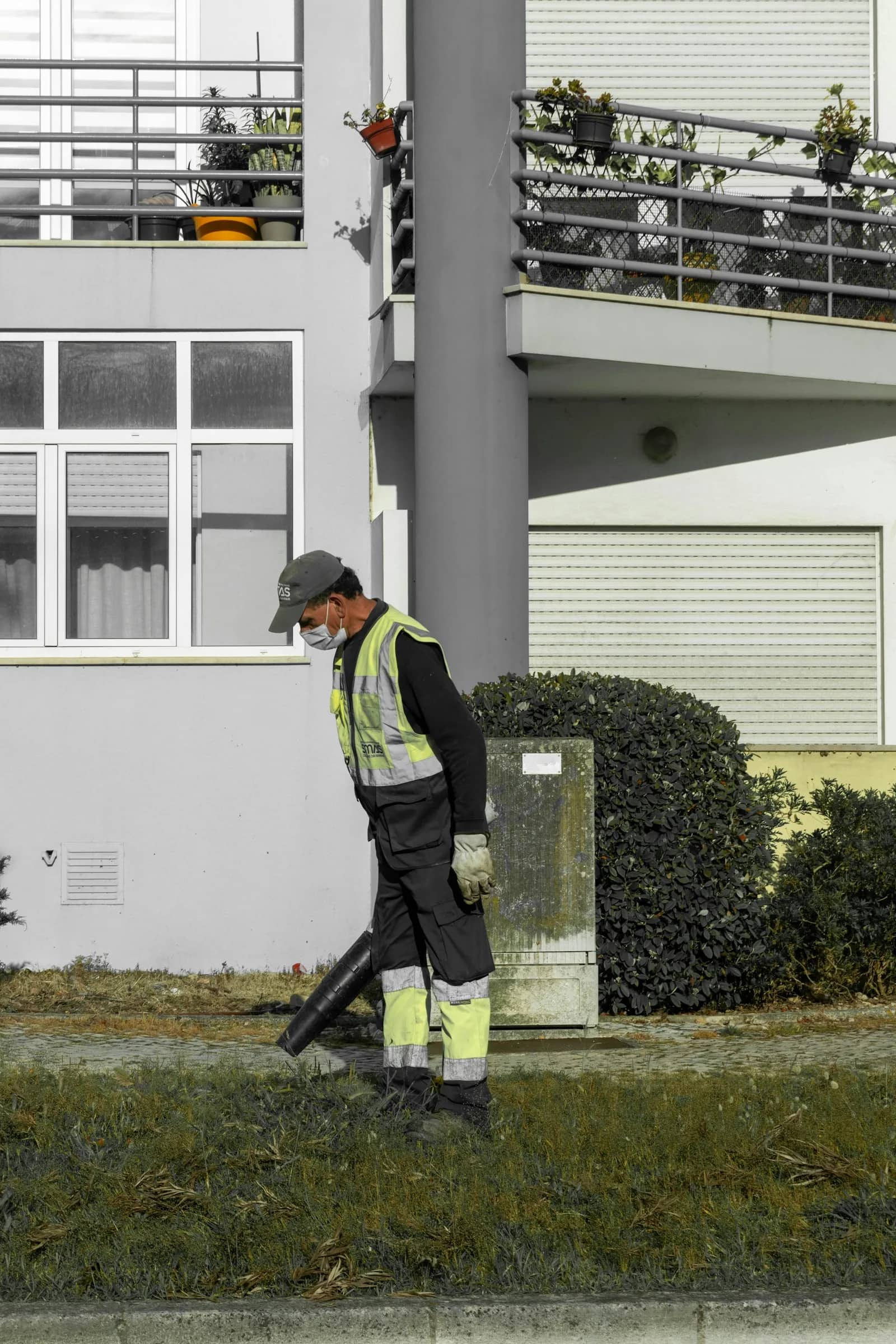 Landscaping and lawn care: a groundskeeper in a hi-vis vest working beside a clipped hedge at the foot of a building, the hedge squared off along the wall behind him.
