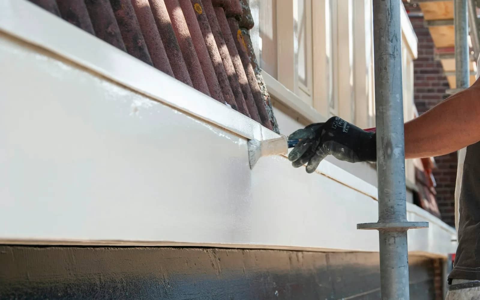 Painting prep: a gloved hand working a brush along the joint where a board meets the trim above it, on a house wrapped in scaffolding.