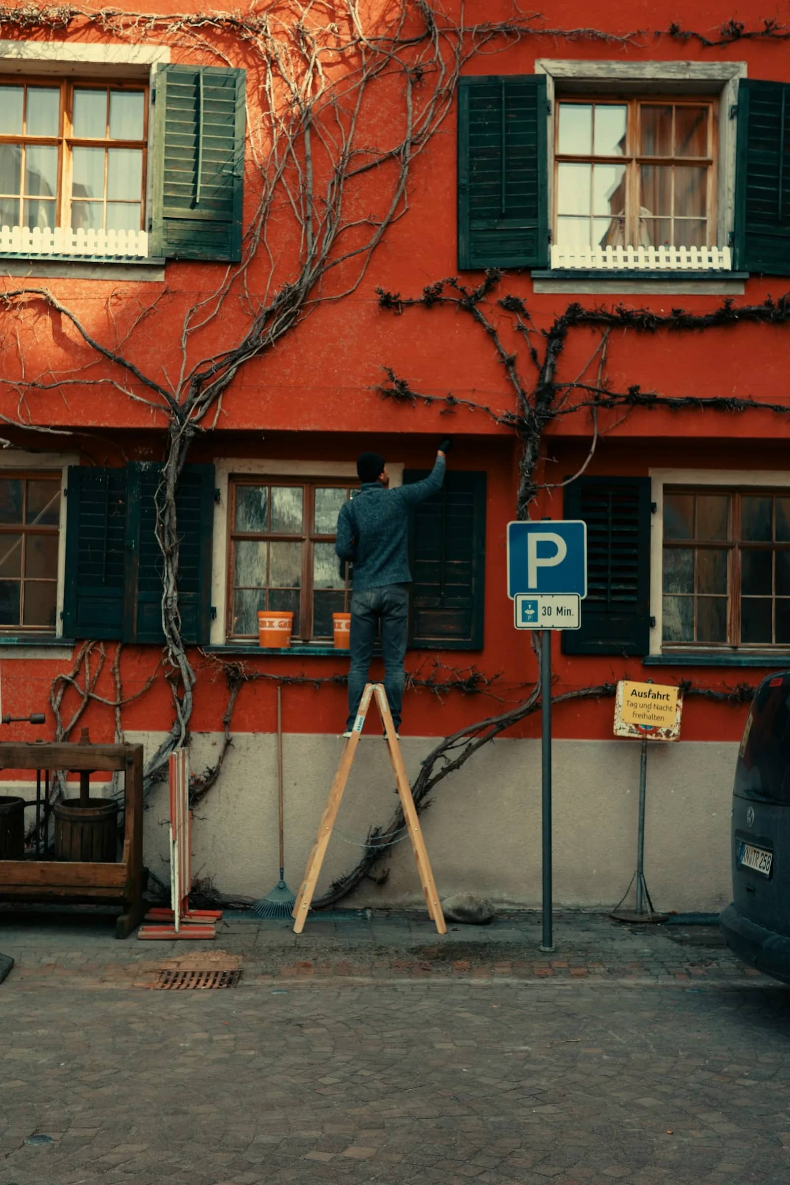 Painting: a decorator on a stepladder reaching up to lay colour along the upper wall of a red building, working past the window heads and shutters.
