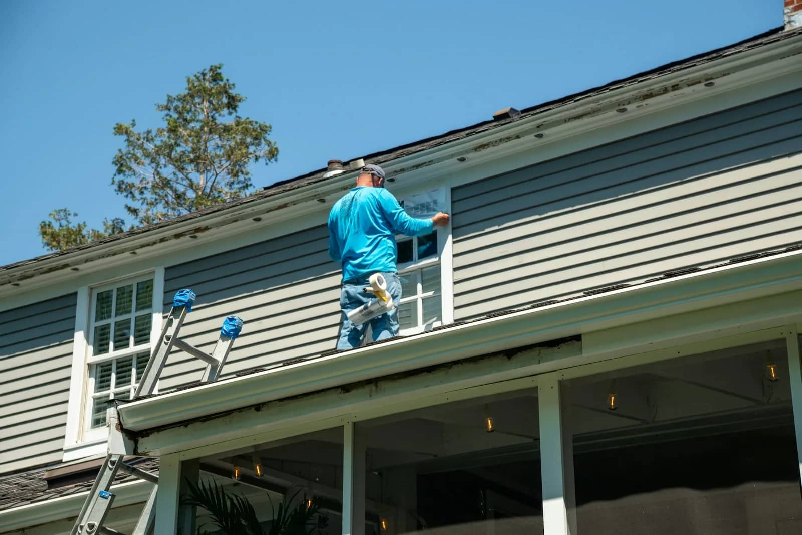 Painting: a decorator on a ladder taping sheeting over a window below the eaves, the covered opening squared off against the siding around it.
