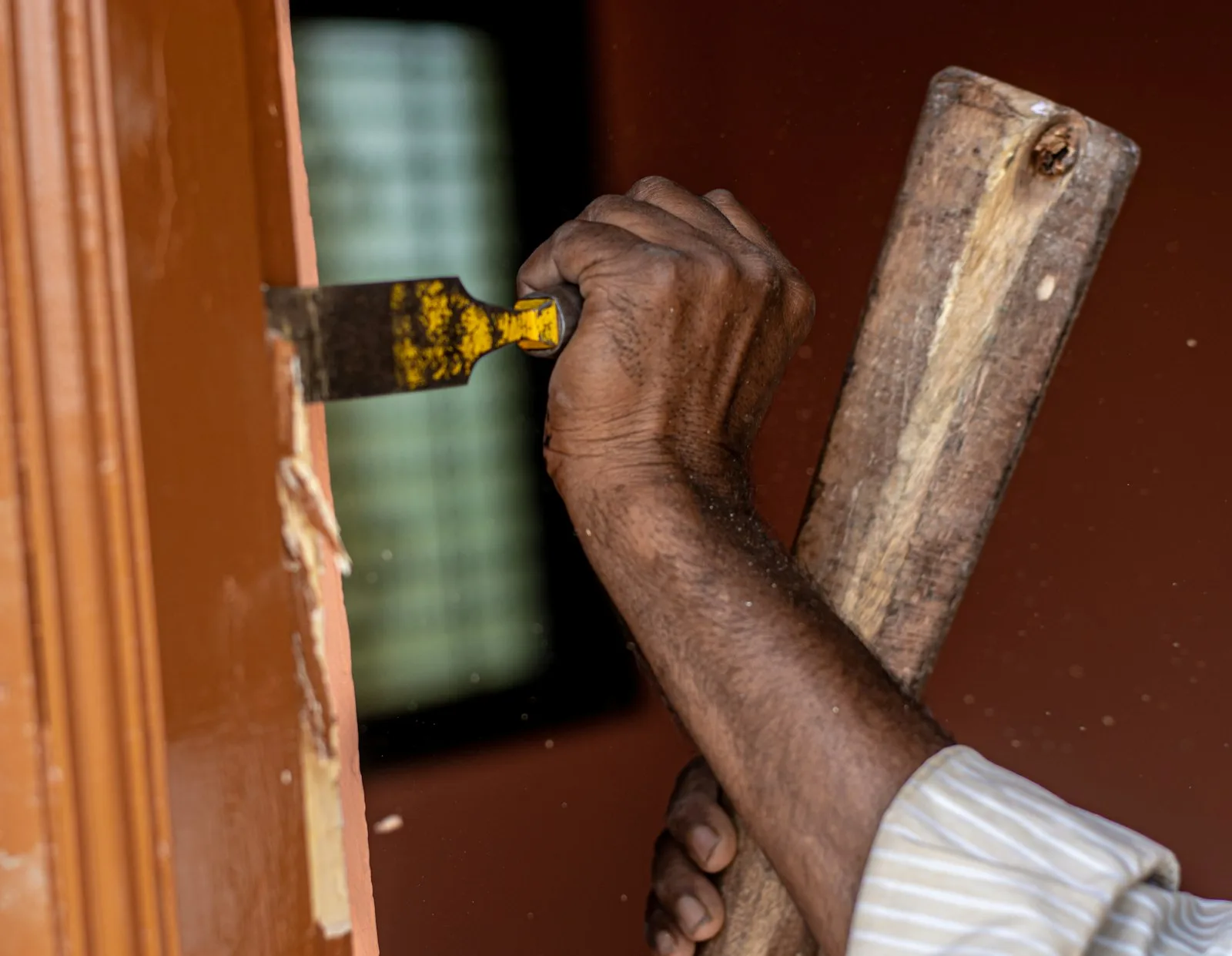 Painting: a hand working a scraper along a painted timber edge, lifting the old finish away in flakes and leaving bare wood behind the blade.