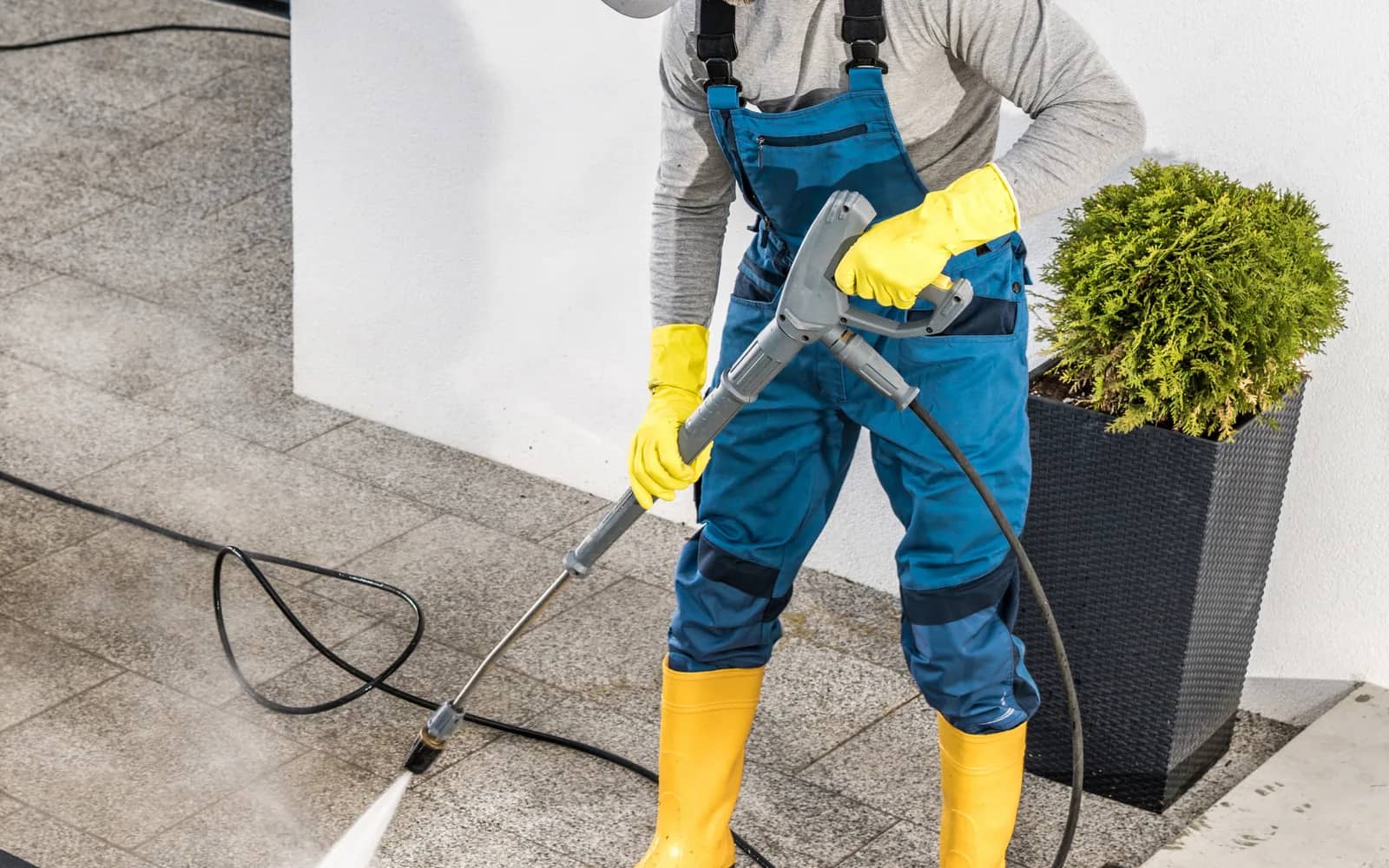 Power washing: an operator in overalls and waterproof boots working a lance across grey paving slabs, the spray driving grime out of the joint line.