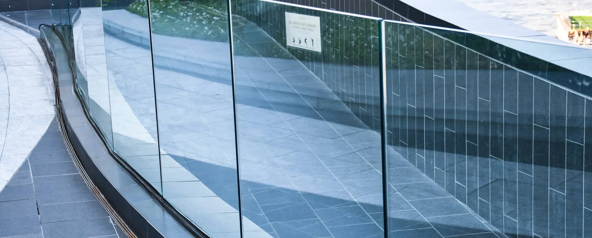 A frameless glass balustrade curving along a paved terrace, the panels clean enough to read the stone paving and the planted bank straight through them.